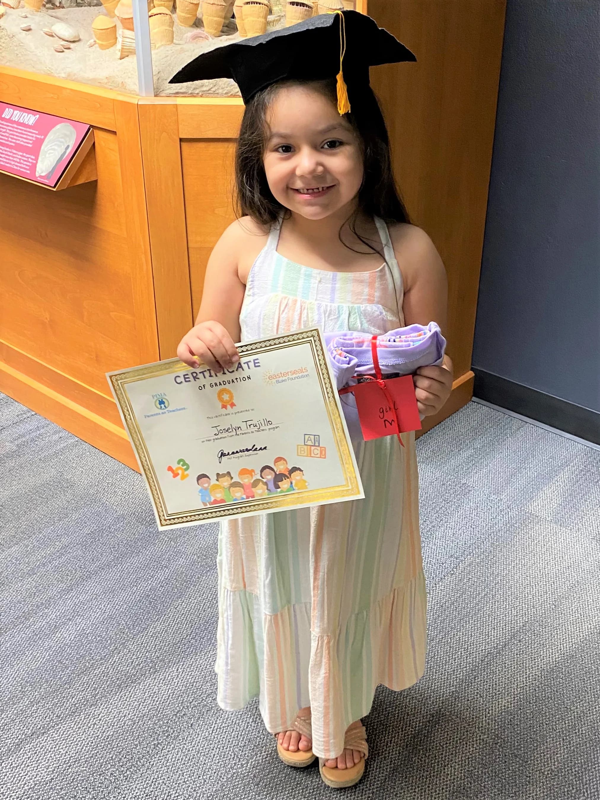 A little girl in a graduation cap holding her certificate and a new pair of pajamas